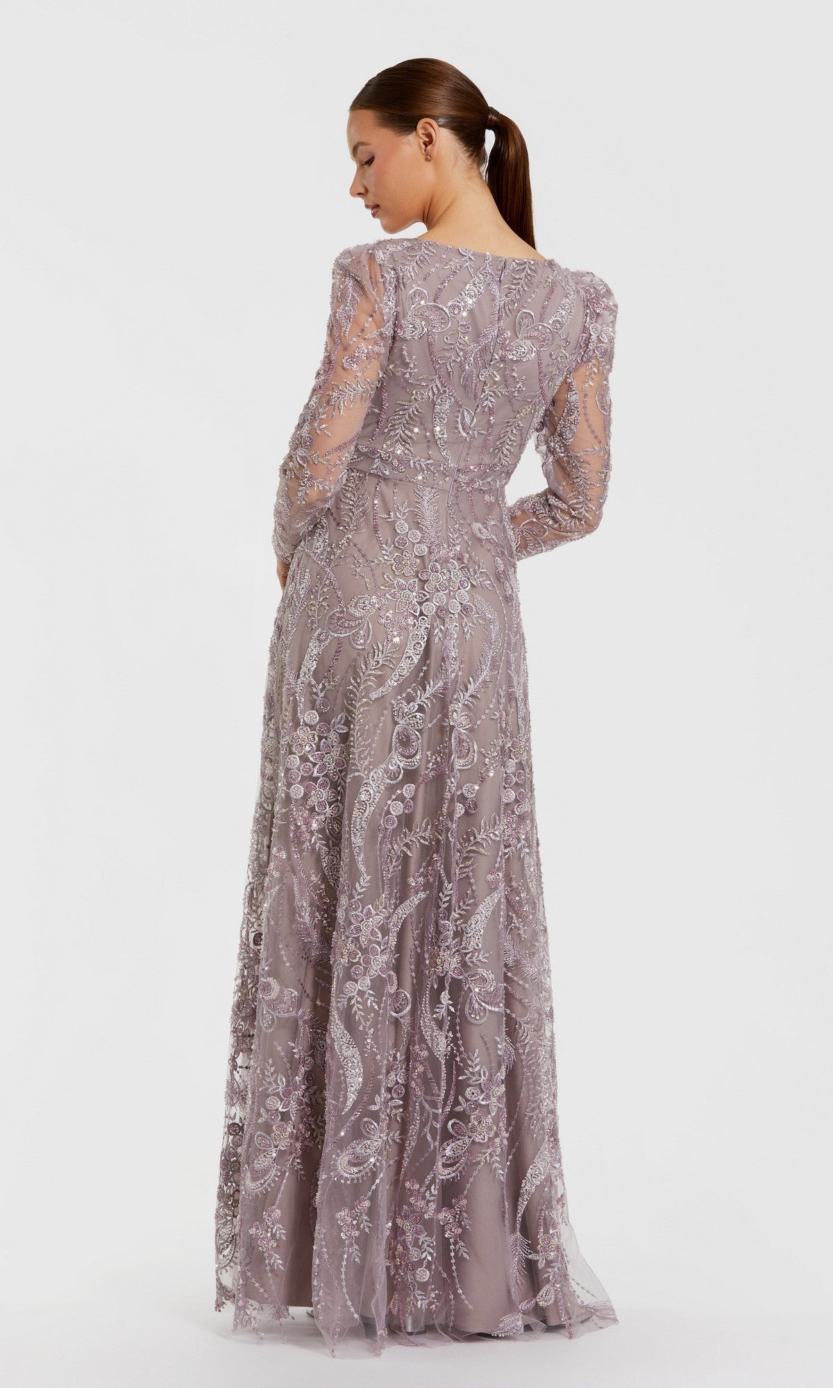 A woman in the Long Formal Dress: Mac Duggal 68908—a sheer, lavender lace gown featuring delicate embroidery—stands with her back to the camera, her hair styled in a low ponytail, against a simple light backdrop.